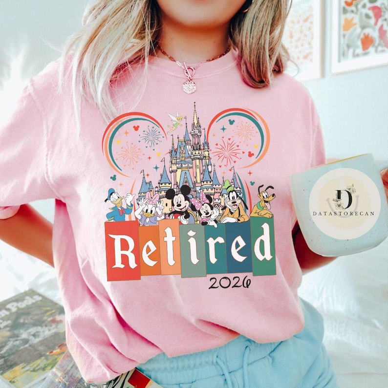 May include: Pink sweatshirt featuring a colorful graphic of a castle, fireworks, and Disney characters. The word "Retired" is displayed in large letters, with the year "2026" below. A light blue mug with the text "DATASTORECAN" is also visible.
