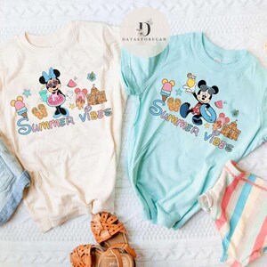 May include: Two light-colored t-shirts with a summer theme. One shirt features Mickey Mouse with the text "Summer Vibes". The other shirt features Minnie Mouse with the text "Summer Vibes".