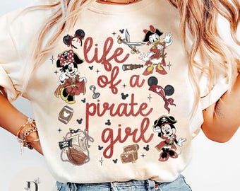 May include: Cream-colored t-shirt featuring the phrase "life of a pirate girl" in red script. The shirt has illustrations of Disney characters dressed as pirates, including Minnie Mouse, Donald Duck, and Mickey Mouse.