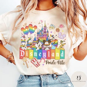 May include: Cream-colored Disneyland Pride Nite t-shirt featuring a colorful graphic. The design includes Mickey and Minnie Mouse, Donald Duck, and other Disney characters, along with a rainbow castle and balloons. The words "Disneyland Pride Nite" are prominently displayed.