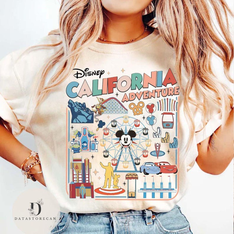 May include: Cream-colored t-shirt featuring a colorful graphic of Disney California Adventure. The design includes the park's name, a Ferris wheel with Mickey Mouse, roller coaster, and other iconic attractions. The shirt is a casual wear item.