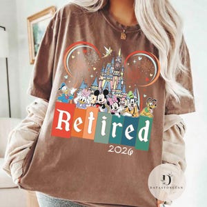 May include: Brown t-shirt with a colorful graphic featuring Disney characters, a castle, fireworks, and the word "Retired" in white letters above the year "2026". The design includes Mickey and Minnie Mouse, Donald Duck, and Goofy.