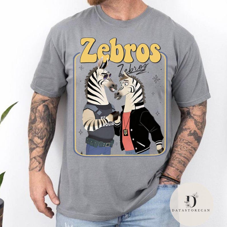 Pu&ograve; includere: T-shirt grigia con un'illustrazione a cartoni animati di due zebre. Una zebra indossa occhiali da sole e un'uniforme della polizia, l'altra una giacca nera. Il testo "Zebros" &egrave; sopra le zebre in giallo.