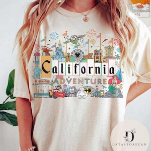 Op de afbeelding: Een wit T-shirt met een kleurrijk grafisch ontwerp met een California Adventure-thema. Het ontwerp bevat iconische personages en attracties uit het pretpark, waaronder Mickey Mouse, Cars en Toy Story.