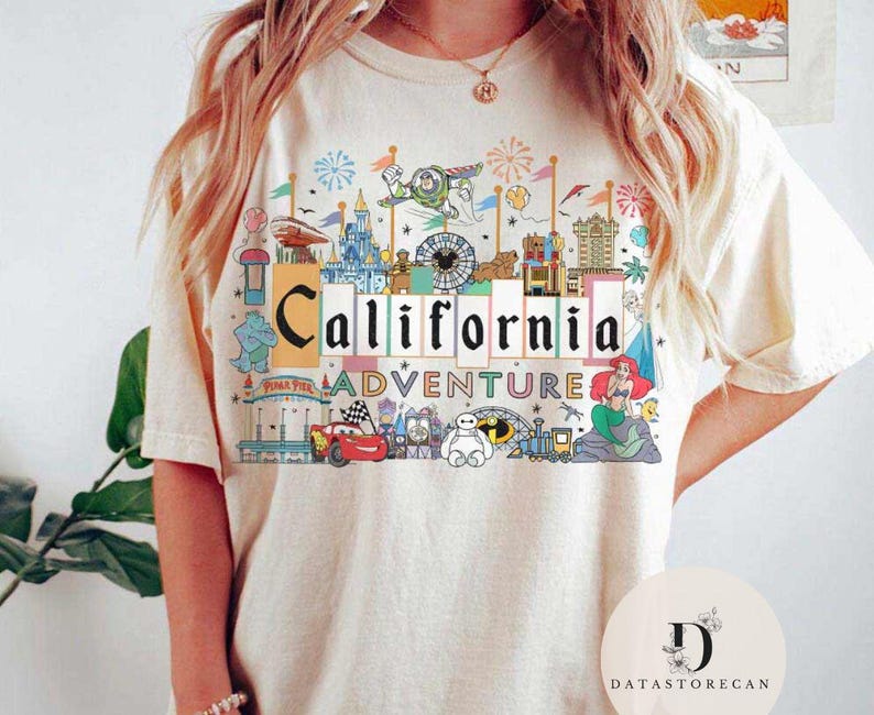Op de afbeelding: Een wit T-shirt met een kleurrijk grafisch ontwerp met een California Adventure-thema. Het ontwerp bevat iconische personages en attracties uit het pretpark, waaronder Mickey Mouse, Cars en Toy Story.
