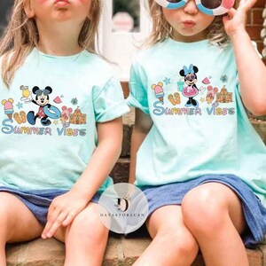 May include: Two children wearing light blue t-shirts with a Mickey Mouse and Minnie Mouse summer theme graphic. The shirts have the text "Summer Vibes" printed on them.