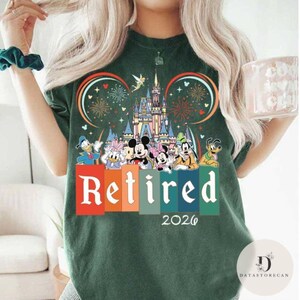 May include: Dark green t-shirt featuring a colorful graphic of Disney characters, a castle, fireworks, and the word "Retired" with the year "2026". The design includes Mickey and Minnie Mouse, Donald Duck, and Goofy.