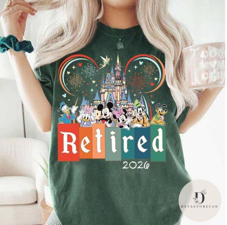 May include: Dark green t-shirt featuring a colorful graphic of Disney characters, a castle, fireworks, and the word "Retired" with the year "2026". The design includes Mickey and Minnie Mouse, Donald Duck, and Goofy.
