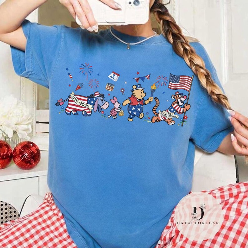 Op de afbeelding: Lichtblauw T-shirt met een patriottisch cartoonontwerp. De afbeelding toont Winnie de Poeh, Knorretje, Iejoor en Teigetje, elk versierd met Amerikaanse vlagmotieven en vuurwerk. Tekst bevat "1776-2026" en "250".