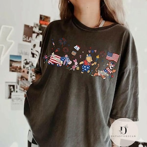 Op de afbeelding: Donkergrijs T-shirt met een patriottisch ontwerp met Winnie de Poeh-personages. De afbeelding bevat een Amerikaanse vlag, vuurwerk en de tekst "1776-2026". De personages zijn gekleed in rood, wit en blauw.