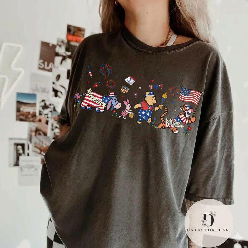 Op de afbeelding: Donkergrijs T-shirt met een patriottisch ontwerp met Winnie de Poeh-personages. De afbeelding bevat een Amerikaanse vlag, vuurwerk en de tekst "1776-2026". De personages zijn gekleed in rood, wit en blauw.