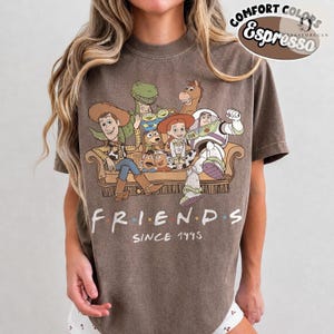 May include: A brown short-sleeve t-shirt featuring a graphic of Toy Story characters sitting on a sofa with the text "FRIENDS SINCE 1995" printed below. The shirt includes a Comfort Colors Espresso label in the upper right corner of the image.