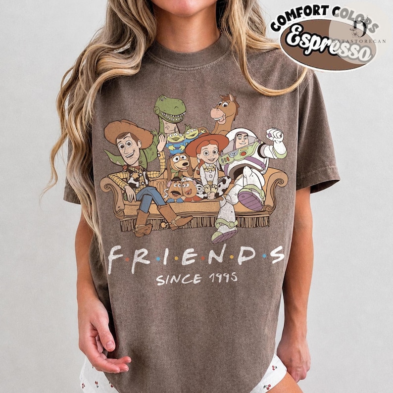 May include: A brown short-sleeve t-shirt featuring a graphic of Toy Story characters sitting on a sofa with the text "FRIENDS SINCE 1995" printed below. The shirt includes a Comfort Colors Espresso label in the upper right corner of the image.