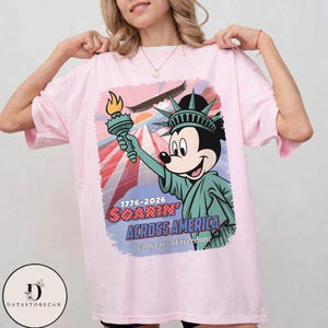 May include: A pale pink t-shirt with a graphic of Mickey Mouse dressed as the Statue of Liberty, holding a torch. The design includes the text "1776-2026 Soarin' Across America" and "250 years of freedom."