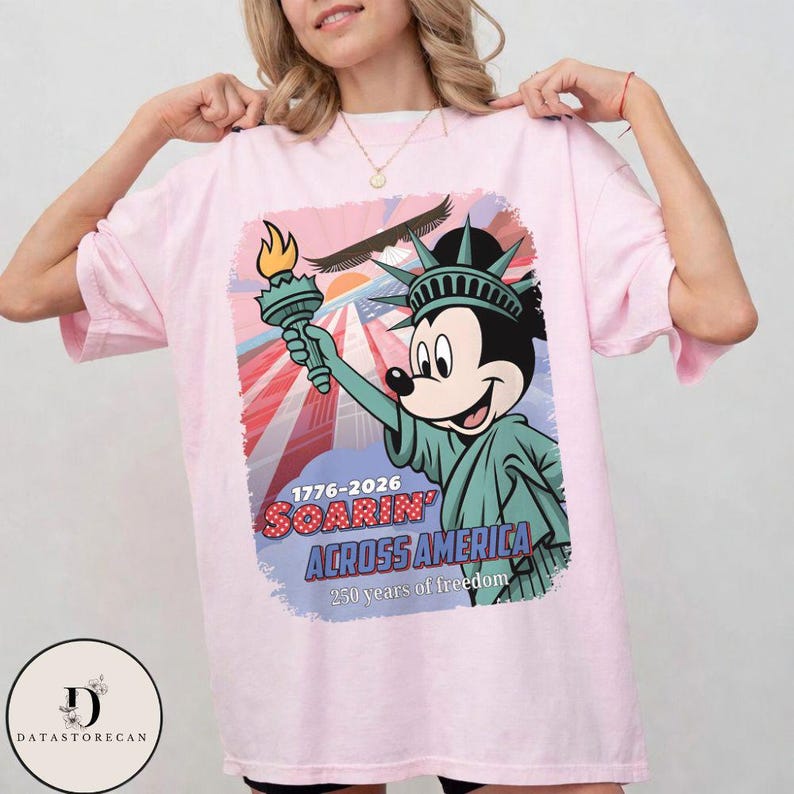 May include: A pale pink t-shirt with a graphic of Mickey Mouse dressed as the Statue of Liberty, holding a torch. The design includes the text "1776-2026 Soarin' Across America" and "250 years of freedom."