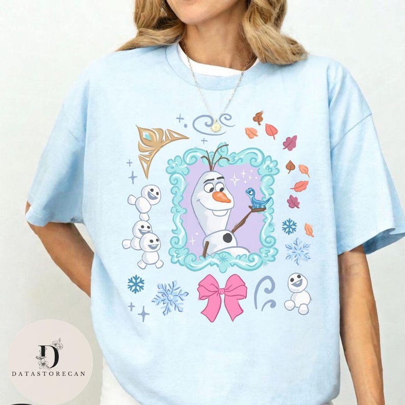 Peut inclure: T-shirt bleu clair avec un motif d'Olaf de La Reine des Neiges, encadr&eacute; dans une bordure d&eacute;corative. Le motif comprend des flocons de neige, des feuilles et un n&oelig;ud rose. Le t-shirt a des manches courtes et un col rond.