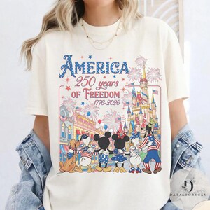 May include: Off-white t-shirt featuring a graphic commemorating 250 years of American freedom. The design includes fireworks, a castle, and Disney characters such as Mickey and Minnie Mouse, Donald Duck, and Goofy.