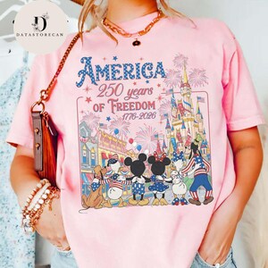 May include: Pale pink t-shirt featuring the text "America 250 years of Freedom 1776-2026" and a graphic of Disney characters in patriotic outfits, with a castle and fireworks in the background.