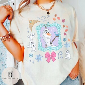 Peut inclure: T-shirt cr&egrave;me avec une illustration encadr&eacute;e d'Olaf de La Reine des Neiges, tenant un oiseau bleu. Le motif comprend des flocons de neige, un n&oelig;ud rose et de petits bonhommes de neige. Le t-shirt a un effet tie-dye.