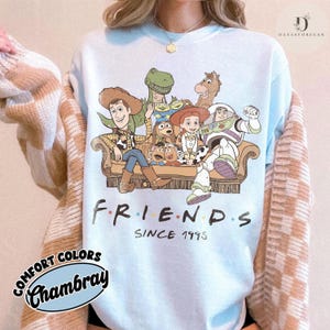May include: A light blue crewneck sweatshirt featuring a graphic print of various animated characters sitting on a sofa. Below the characters, the text reads FRIENDS SINCE 1995. The sweatshirt is worn under a beige and white checkered cardigan sweater.