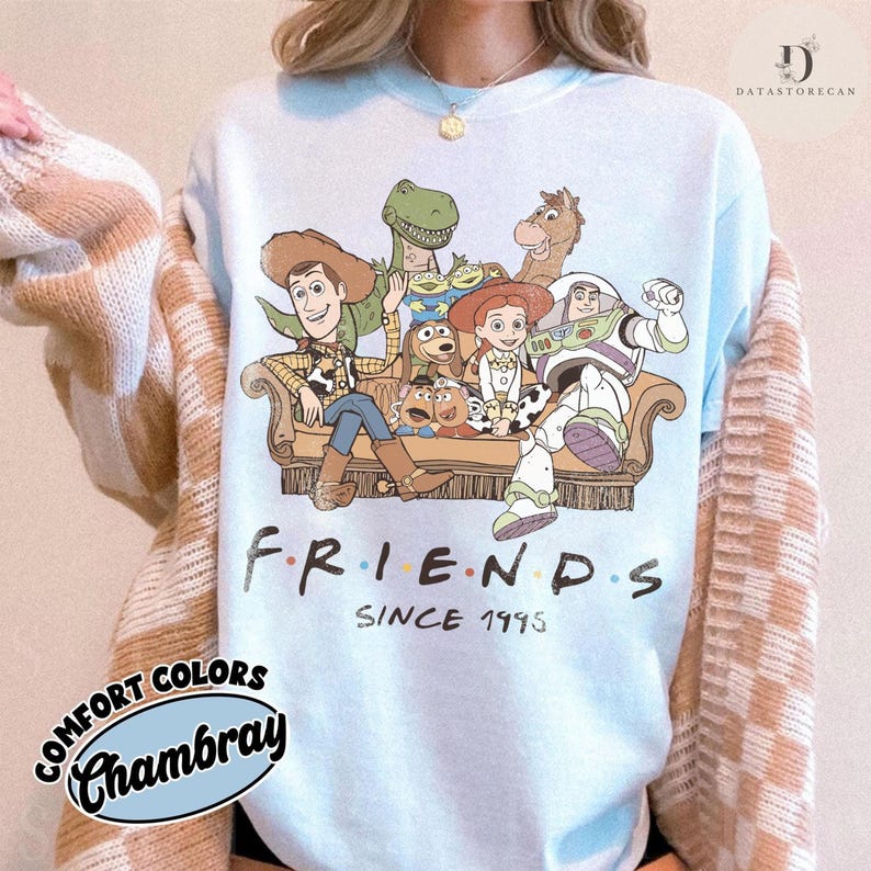 May include: A light blue crewneck sweatshirt featuring a graphic print of various animated characters sitting on a sofa. Below the characters, the text reads FRIENDS SINCE 1995. The sweatshirt is worn under a beige and white checkered cardigan sweater.