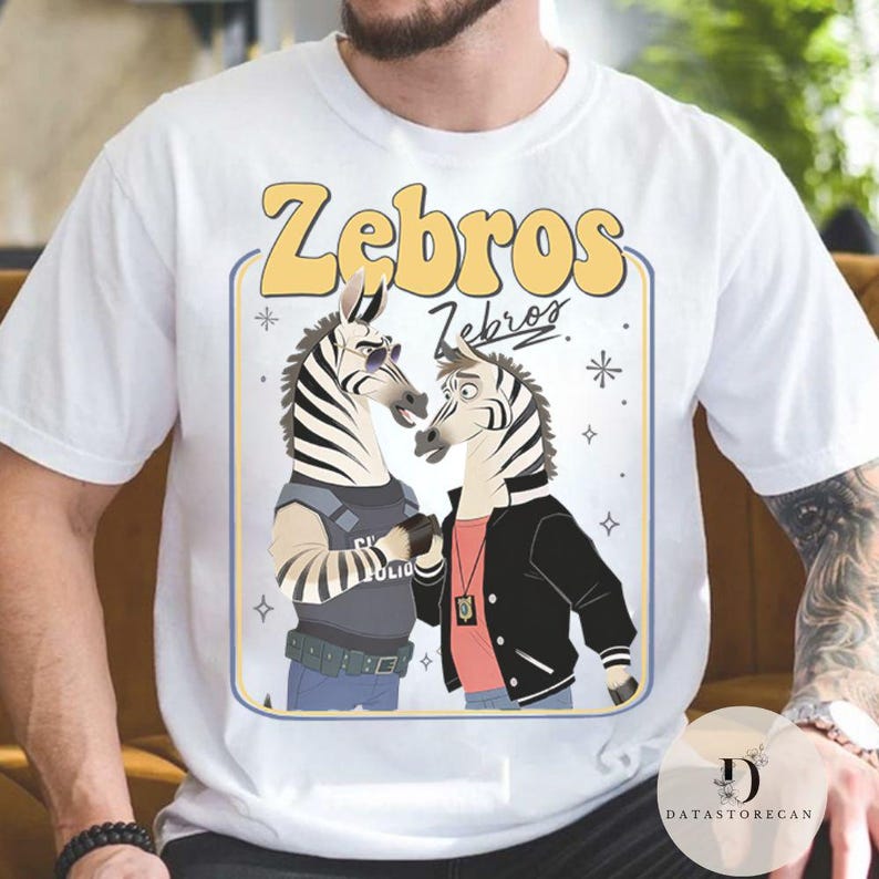 Pu&ograve; includere: T-shirt bianca con una grafica a cartone animato di due zebre. Le zebre sono antropomorfe, con occhiali da sole e una giacca. La maglietta ha la scritta "Zebros" in giallo, con un design retr&ograve;.