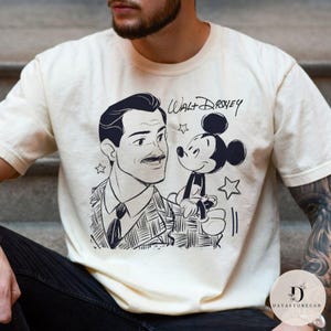 May include: Cream-colored t-shirt featuring a black and white illustration of Walt Disney and Mickey Mouse. The text "Walt Disney" is written above the characters. The shirt has a classic crew neck and short sleeves.