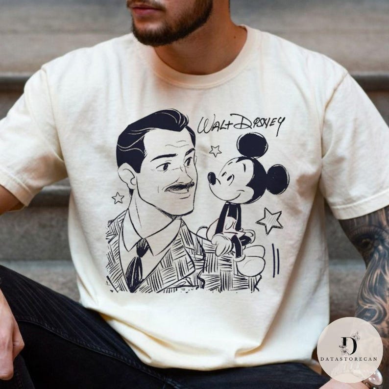 May include: Cream-colored t-shirt featuring a black and white illustration of Walt Disney and Mickey Mouse. The text "Walt Disney" is written above the characters. The shirt has a classic crew neck and short sleeves.