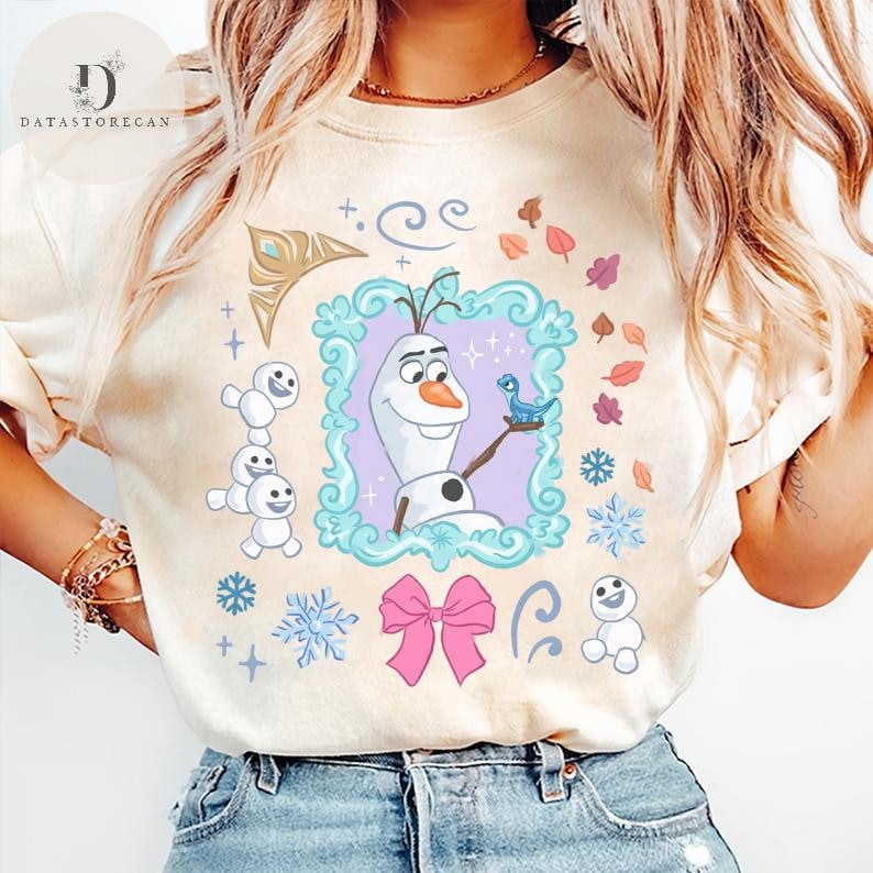 Peut inclure: T-shirt beige avec une image de dessin anim&eacute; d'Olaf de La Reine des Neiges. Olaf est dans un cadre d&eacute;coratif avec une bordure bleue, tenant un oiseau bleu. Le t-shirt a &eacute;galement des flocons de neige, un n&oelig;ud rose et d'autres &eacute;l&eacute;ments d&eacute;coratifs.