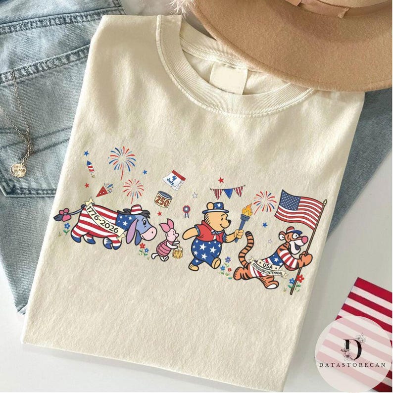 Op de afbeelding: Cr&egrave;mekleurig T-shirt met een patriottisch ontwerp met stripfiguren. Het ontwerp bevat Winnie de Poeh, Knorretje, Iejoor en Tijger, elk versierd met kleding in Amerikaanse vlagstijl en met patriottische symbolen.