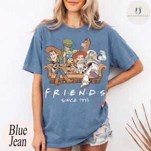 May include: A blue short-sleeve t-shirt featuring a graphic print of various animated characters sitting on a couch. Below the characters, the text reads FRIENDS SINCE 1995 in a white font with colored dots between the letters of the word FRIENDS.
