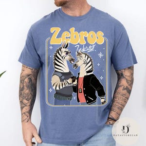 Pu&ograve; includere: T-shirt blu con un'illustrazione a cartoni animati di due zebre. Una zebra &egrave; vestita da poliziotto, l'altra con una giacca. Il testo giallo sopra dice "Zebros".