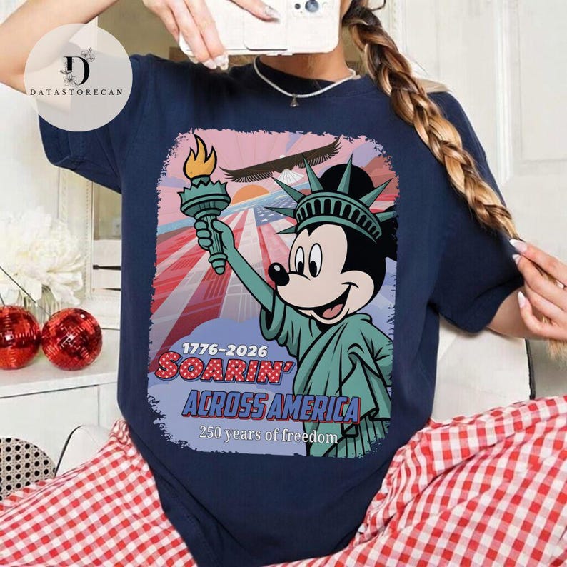 May include: Navy blue t-shirt with a graphic of Mickey Mouse as the Statue of Liberty, holding a torch. The design includes the text "1776-2026 Soarin' Across America" and "250 years of freedom" with a patriotic backdrop.