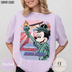 May include: Orchid-coloured t-shirt with a graphic of Mickey Mouse as the Statue of Liberty, holding a torch. The design includes the text "SOARIN' ACROSS AMERICA 1776-2026" and "250 years of freedom."