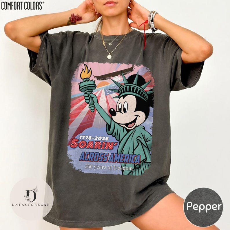 May include: Dark grey t-shirt with a graphic of Mickey Mouse as the Statue of Liberty, holding a torch. The design includes the text "1776-2026 Soarin' Across America" and "250 years of freedom". The shirt is labeled "Pepper".