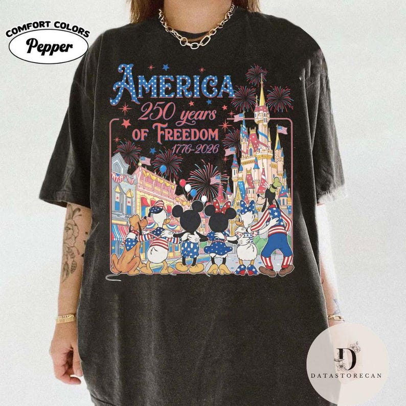 May include: Dark grey t-shirt with a colourful graphic celebrating America's 250 years of freedom. The design features fireworks, a castle, and Disney characters in patriotic attire. The text "AMERICA 250 years of Freedom 1776-2026" is displayed.