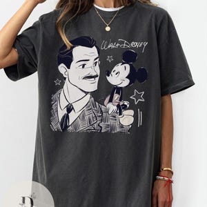 May include: Dark gray t-shirt featuring a black and white illustration of Walt Disney with Mickey Mouse. The shirt has the text "Walt Disney" in a handwritten style above the cartoon characters. The shirt is a relaxed fit.