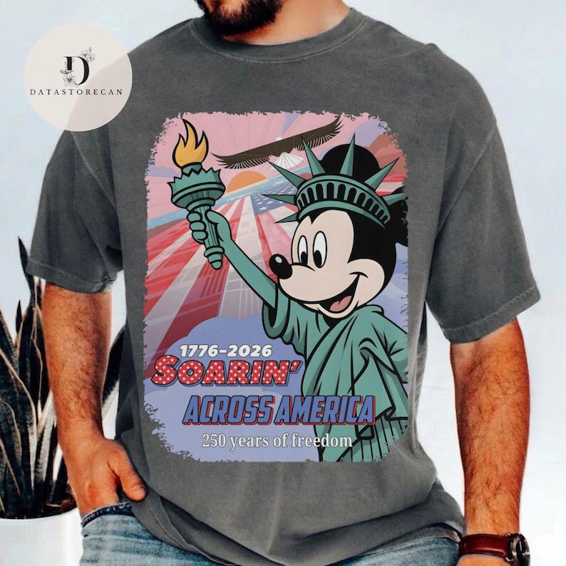 May include: Dark grey t-shirt with a graphic of Mickey Mouse as the Statue of Liberty, holding a torch. The design includes the text "1776-2026 Soarin' Across America" and "250 years of freedom" with a patriotic background.