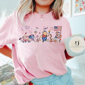 Op de afbeelding: Lichtroze T-shirt met een patriottisch cartoonontwerp. Het ontwerp toont Winnie de Poeh, Knorretje, Iejoor en Tijger in rood, wit en blauw, ter viering van de 250e verjaardag van de VS. Het shirt heeft een ronde hals.