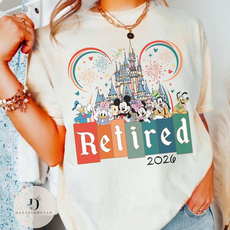 May include: Cream-colored t-shirt featuring a colorful graphic of Disney characters, a castle, fireworks, and the word "Retired" with the year "2026". The design includes Mickey and Minnie Mouse, Donald and Daisy Duck, Goofy, and Pluto.