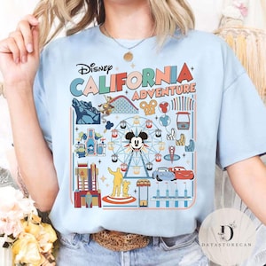 May include: Light blue t-shirt featuring the text "Disney CALIFORNIA ADVENTURE" with colorful graphics of a Ferris wheel, roller coaster, and other park attractions. The shirt has a crew neck and short sleeves.