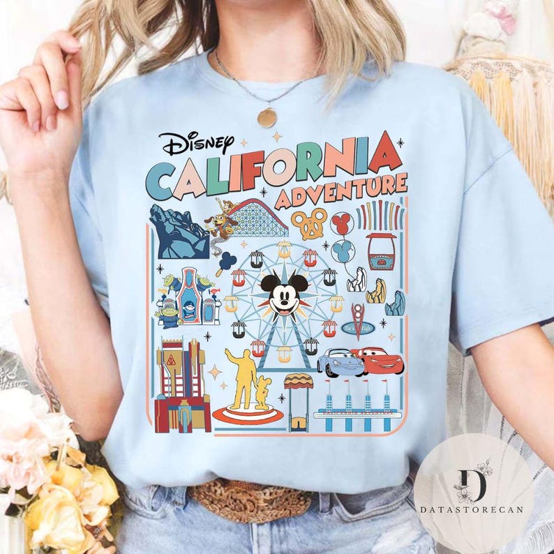 May include: Light blue t-shirt featuring the text "Disney CALIFORNIA ADVENTURE" with colorful graphics of a Ferris wheel, roller coaster, and other park attractions. The shirt has a crew neck and short sleeves.