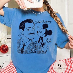 May include: A light blue t-shirt featuring a black illustration of Walt Disney and Mickey Mouse. The text "Walt Disney" is written above the illustration. The shirt has a crew neck and short sleeves.