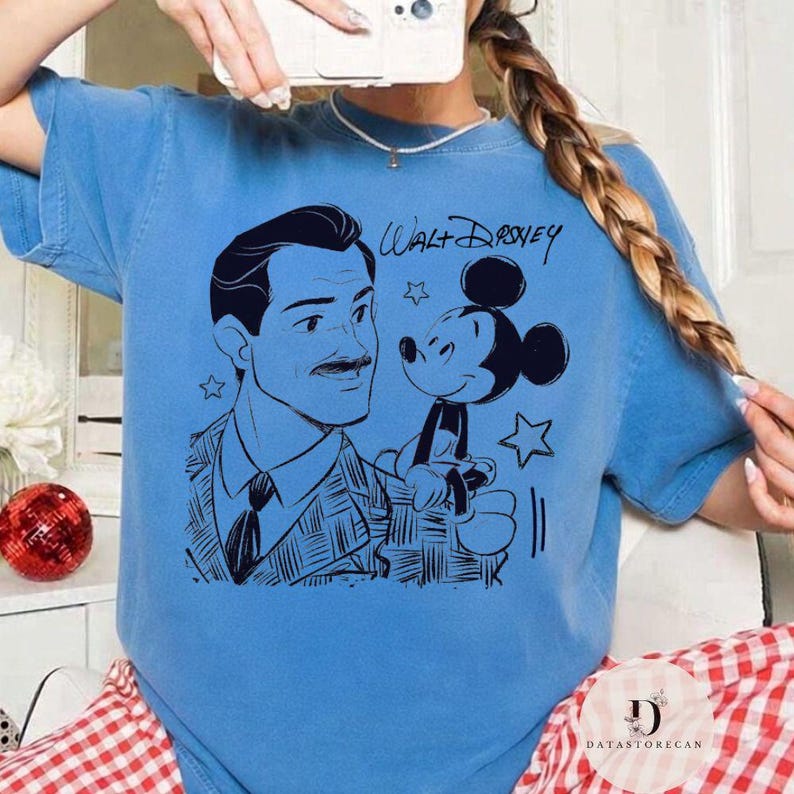 May include: A light blue t-shirt featuring a black illustration of Walt Disney and Mickey Mouse. The text "Walt Disney" is written above the illustration. The shirt has a crew neck and short sleeves.