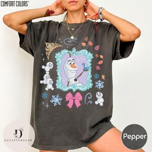 Peut inclure: T-shirt Comfort Colors gris fonc&eacute; avec un motif color&eacute; d'Olaf de La Reine des Neiges. Le motif comprend des flocons de neige, un n&oelig;ud rose et le texte "Pepper". Le t-shirt a une coupe d&eacute;contract&eacute;e.