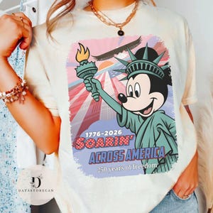 May include: A cream-coloured t-shirt with a graphic of Mickey Mouse as the Statue of Liberty, holding a torch. The design includes the text "1776-2026 Soarin' Across America" and "250 years of freedom."