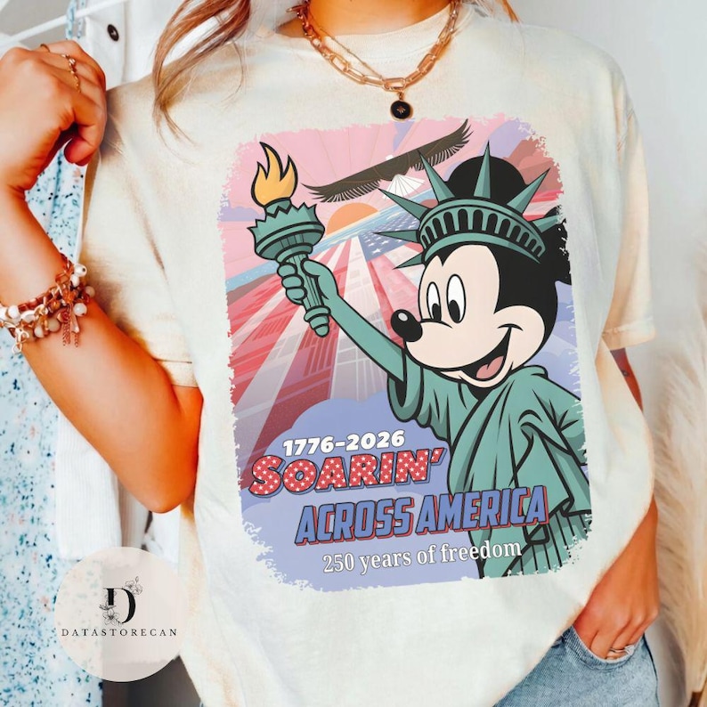 May include: A cream-coloured t-shirt with a graphic of Mickey Mouse as the Statue of Liberty, holding a torch. The design includes the text "1776-2026 Soarin' Across America" and "250 years of freedom."