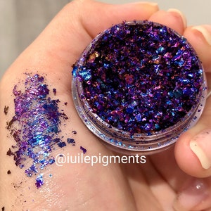 May include: A small jar of iridescent purple and blue glitter flakes. The glitter is loose and has a metallic sheen. The jar is labeled with the text "@iuiilepigments".
