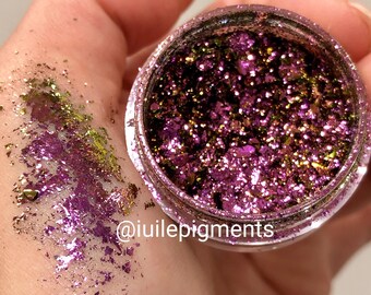May include: A small jar of iridescent purple and green glitter flakes. The glitter is loose and has a metallic sheen. The jar is labeled with the text "@iuiilepigments".