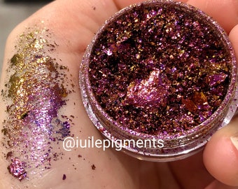 May include: A small jar of iridescent purple and gold glitter flakes. The glitter is loose and has a metallic sheen. The jar is labeled with the text "@iuiilepigments".